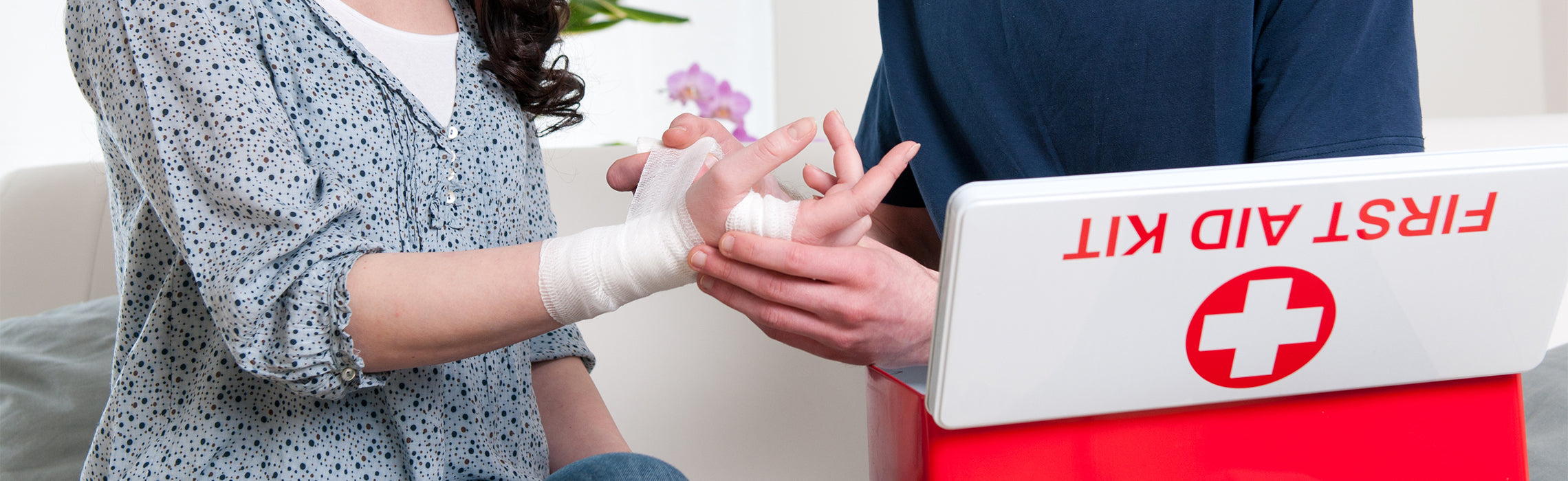 Close-up of a person’s hand being wrapped with a white bandage by another individual next to a kit labeled “FIRST AID KIT,” representing injury care, first aid treatment, and medical support.