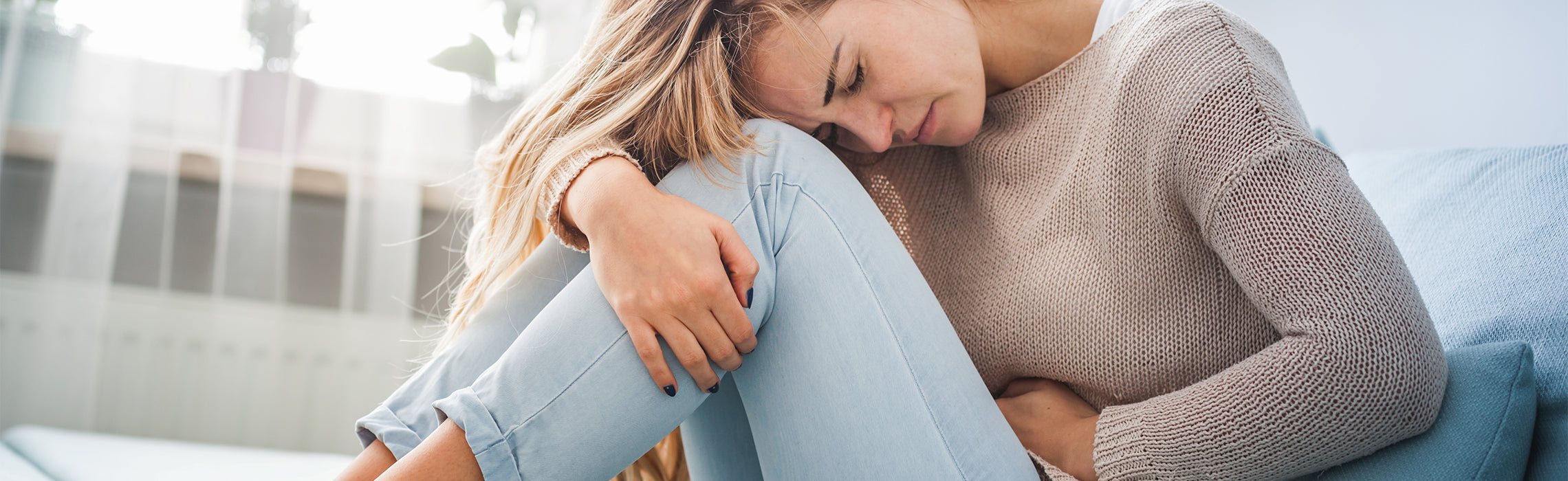 Person sitting curled on a couch holding the lower abdomen with one arm and resting the head on the knees, representing stomach pain, cramps, or discomfort in a home setting with soft natural light.