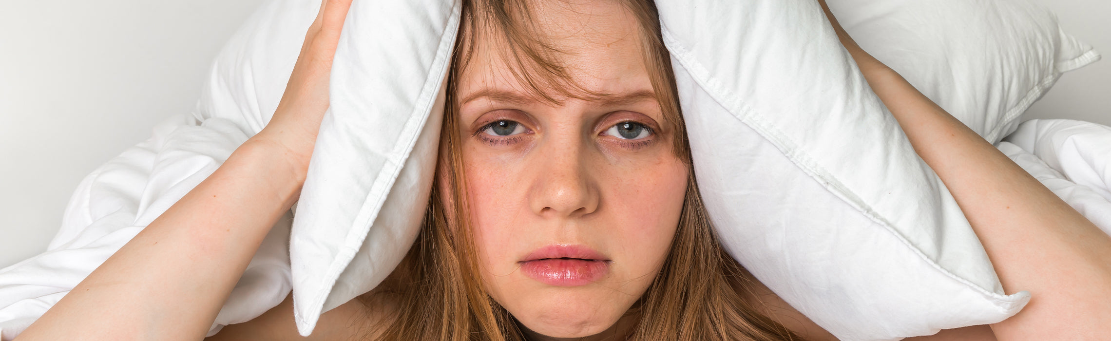 Close-up of a person lying in bed holding a pillow tightly over both ears on white bedding, representing difficulty sleeping, noise disturbance, and insomnia.