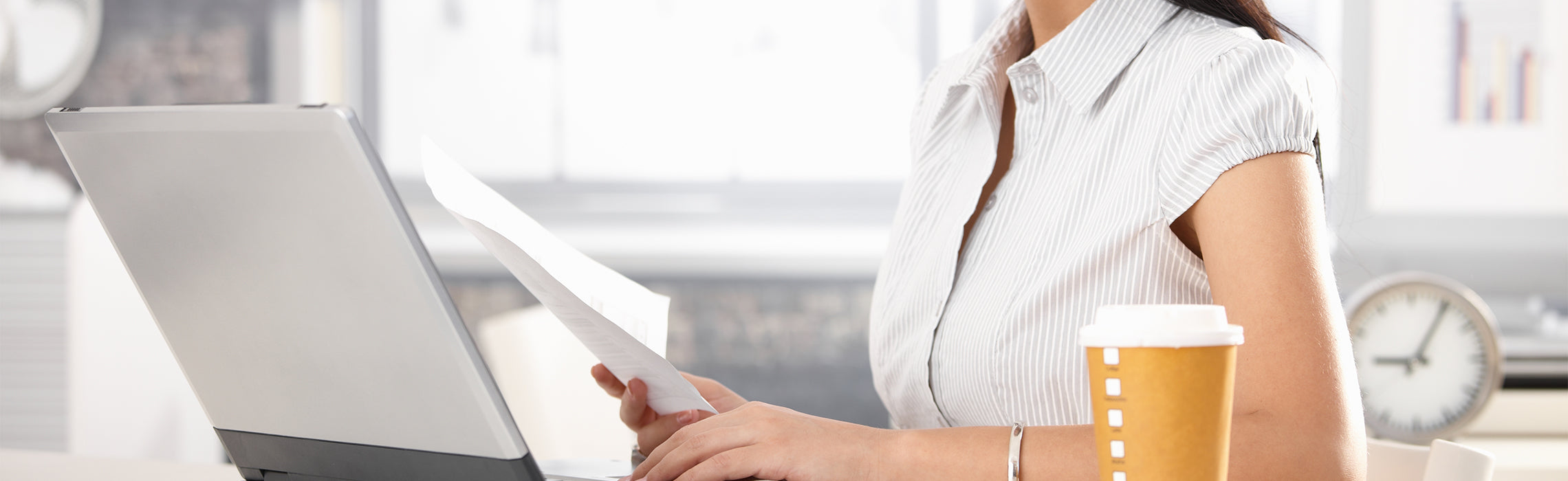 Person seated at a desk reviewing a document beside an open laptop and a takeaway coffee cup, representing office work, productivity, and daily business tasks.