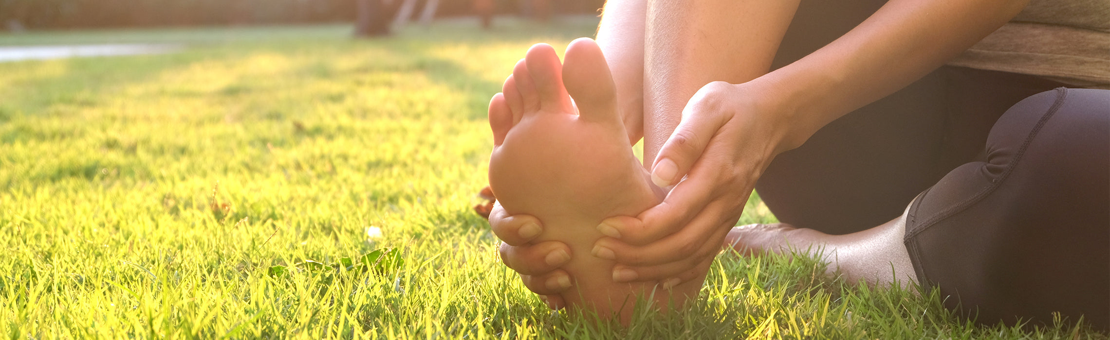 Person sitting on grass outdoors holding their foot in discomfort, suggesting foot pain, soreness, or plantar fasciitis after exercise.