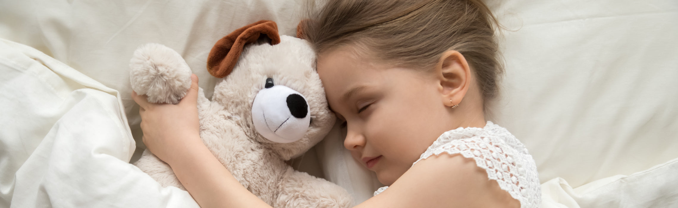 Child lying in bed hugging a stuffed teddy bear while resting on a white pillow and bedding, representing comfort, security, and peaceful sleep.