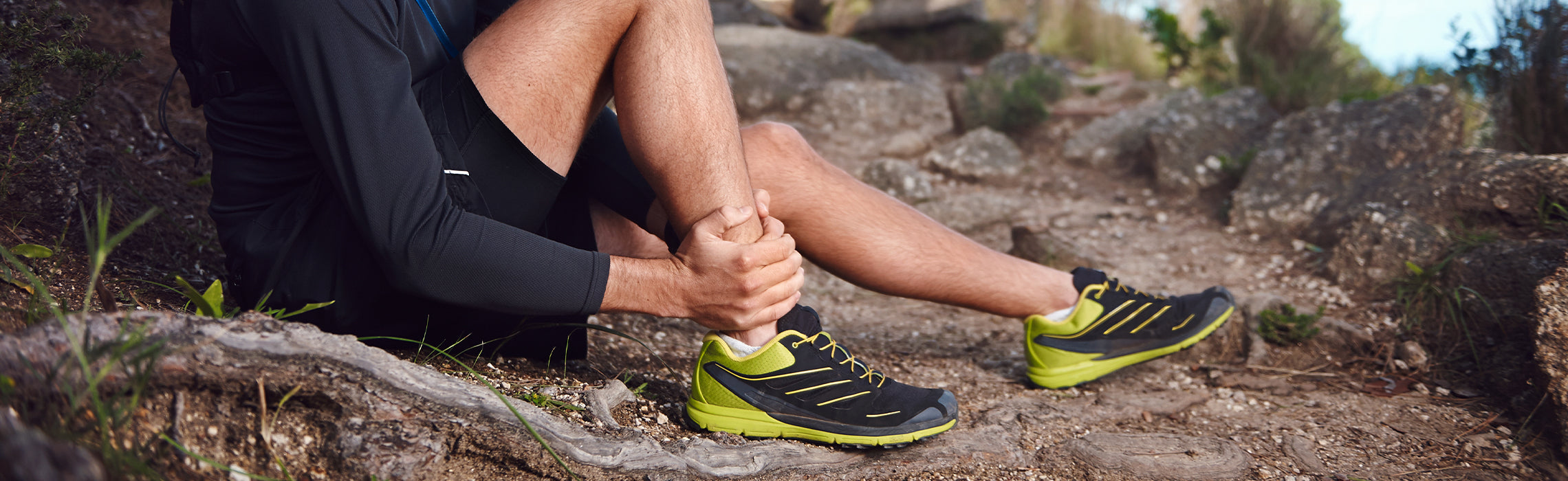Person sitting on a rocky trail holding the ankle with both hands while wearing black and neon running shoes, representing ankle pain, hiking injury, and outdoor exercise strain.