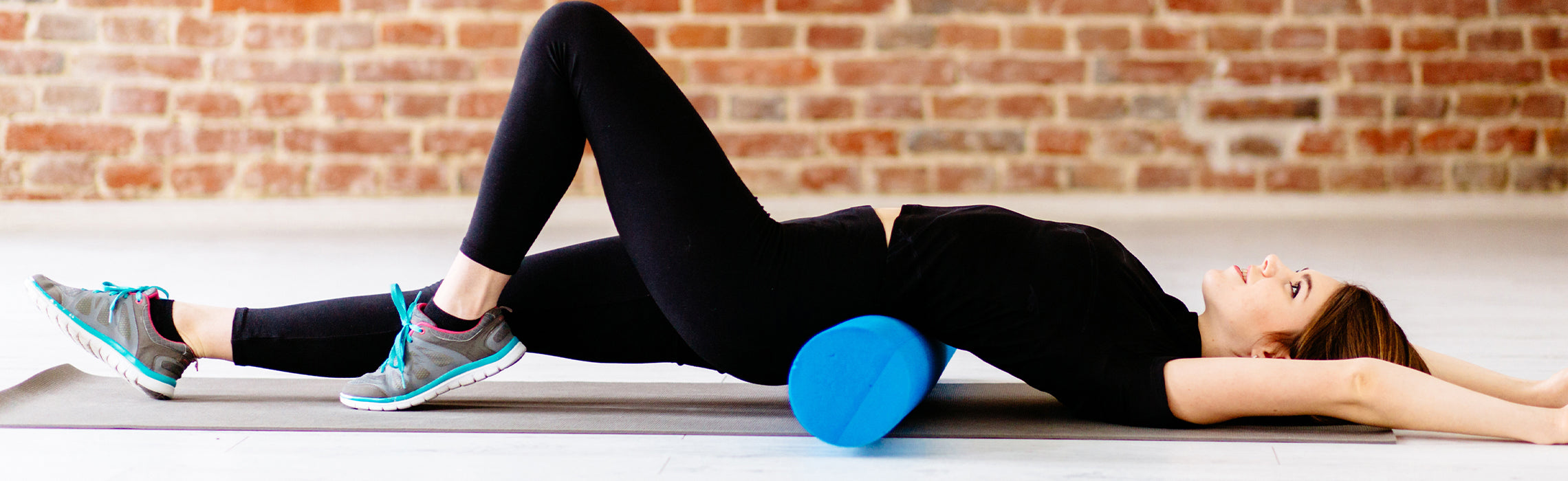 Person lying on a yoga mat using a blue exercise ball under the lower back while performing a stretch, wearing black athletic clothing and running shoes, representing core strengthening, back support, and physical therapy exercise.