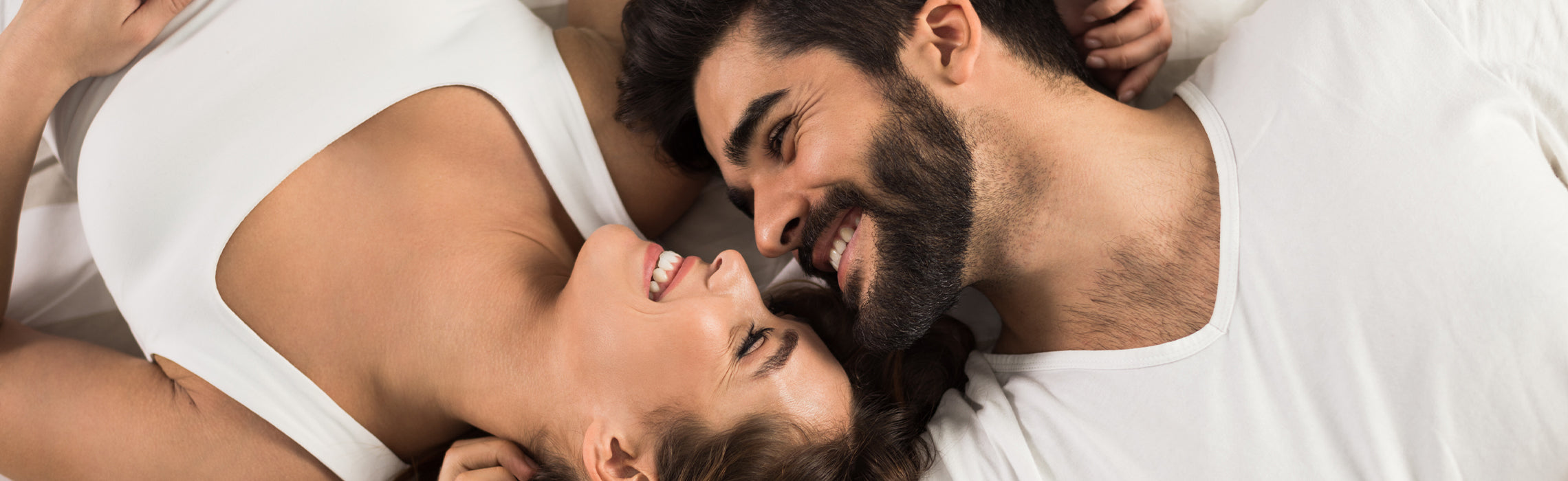 Two people lying on a bed facing each other, smiling closely with heads together on white bedding, representing comfort, connection, and relaxation in a calm bedroom setting.