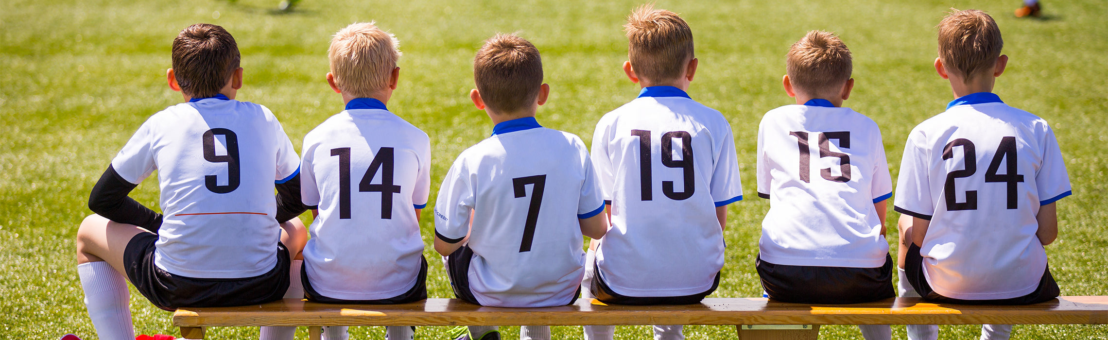 Six youth soccer players sitting on a bench facing a grassy field, wearing white jerseys with visible numbers “9,” “14,” “7,” “19,” “16,” and “24,” representing team sports, youth soccer, and group athletics.