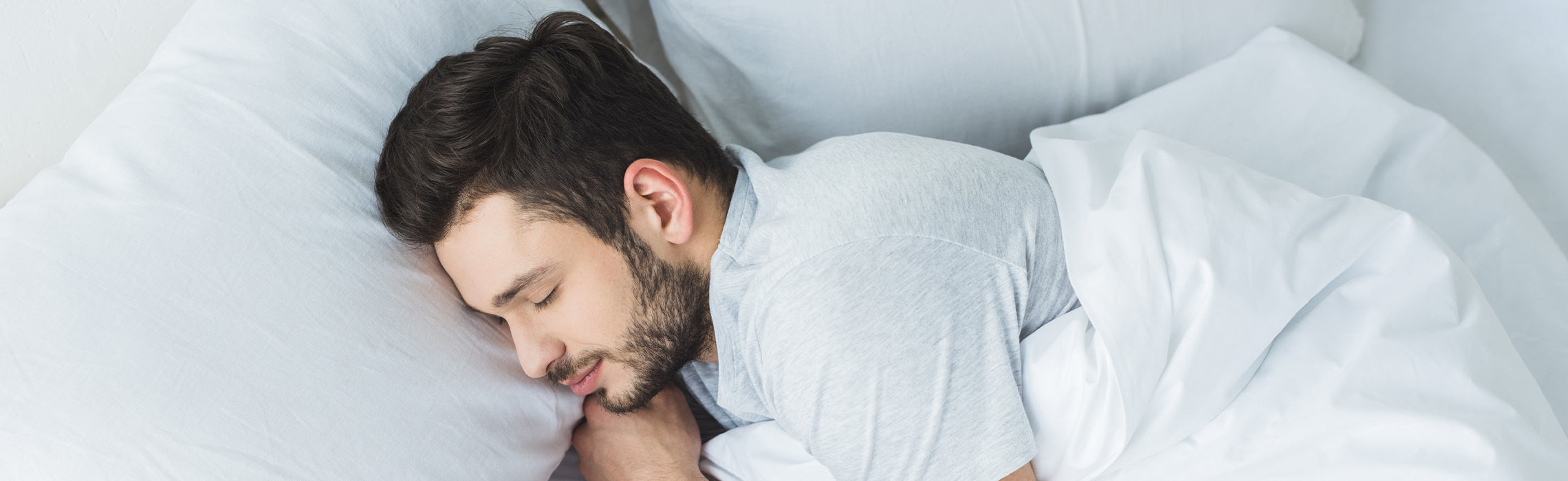 Person lying on a bed on a white pillow and bedding in a side sleeping position with eyes closed, representing restful sleep, comfort, and healthy sleep habits.