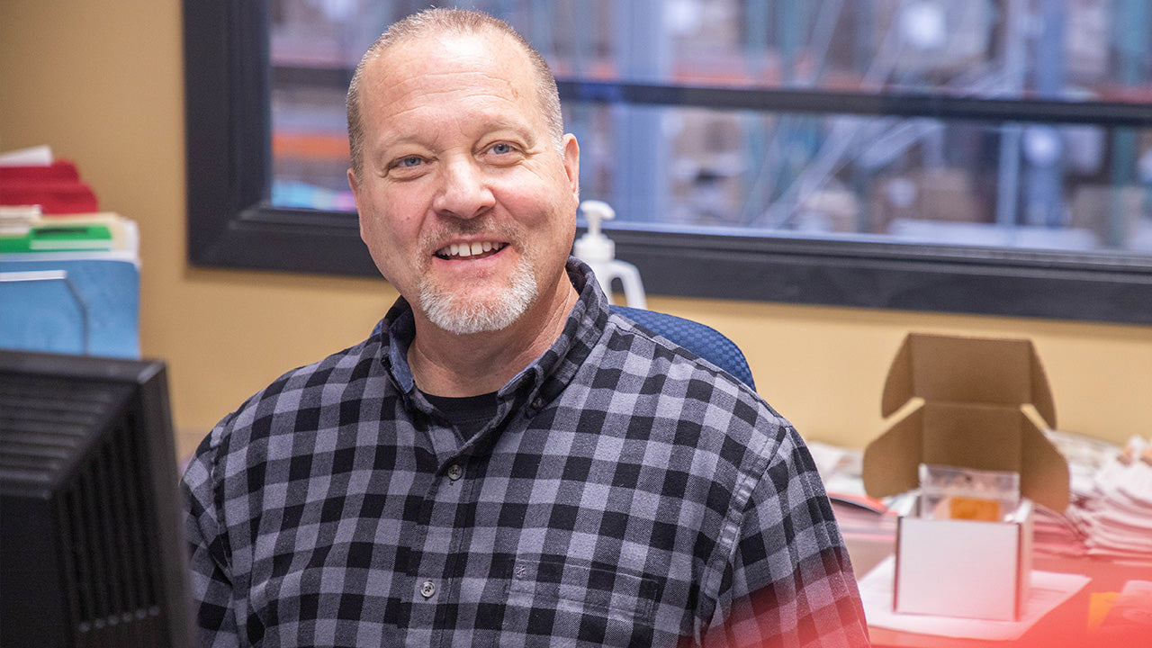 Person seated at a desk in an office, smiling toward the camera, wearing a plaid shirt with a computer, files, and a windowed workspace in the background. No visible text or branding.