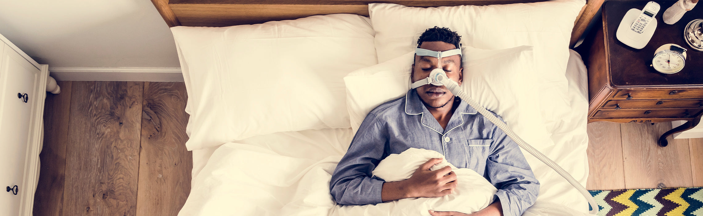Top-down view of a person lying in bed wearing a CPAP mask connected to a machine, resting on white pillows and bedding, with a bedside table holding a phone and clock, representing sleep apnea treatment, breathing support, and improved sleep health.
