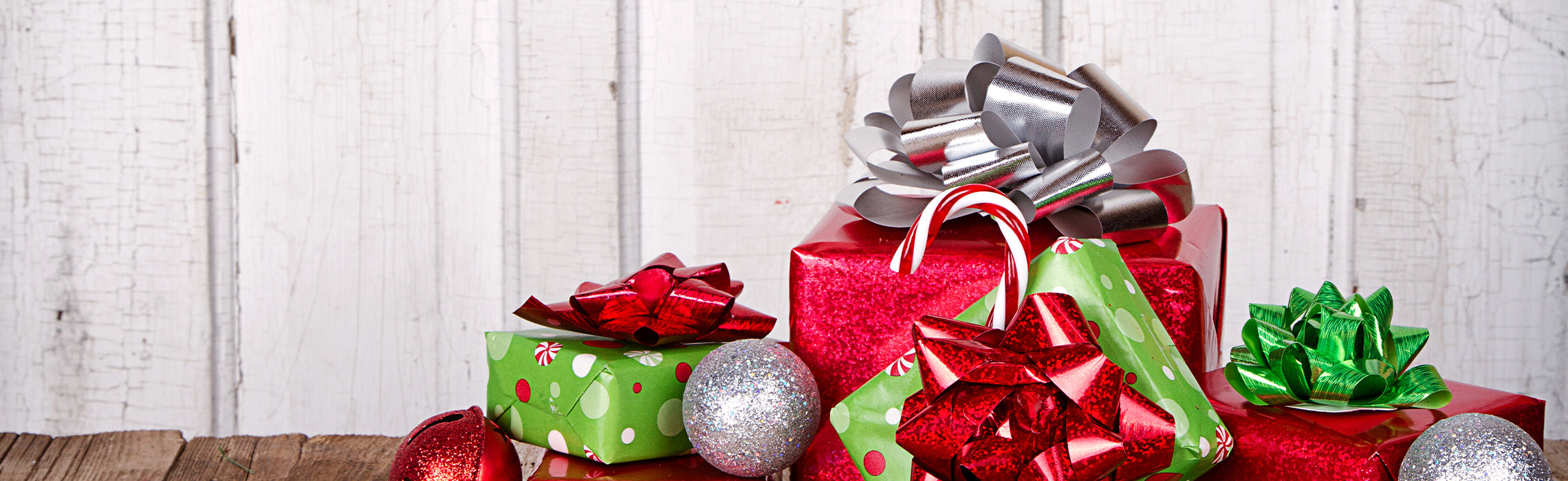 Stack of wrapped Christmas presents in red and green with shiny bows, candy canes, and ornaments arranged on a wooden surface against a white rustic background, representing holiday gifting and festive decor.