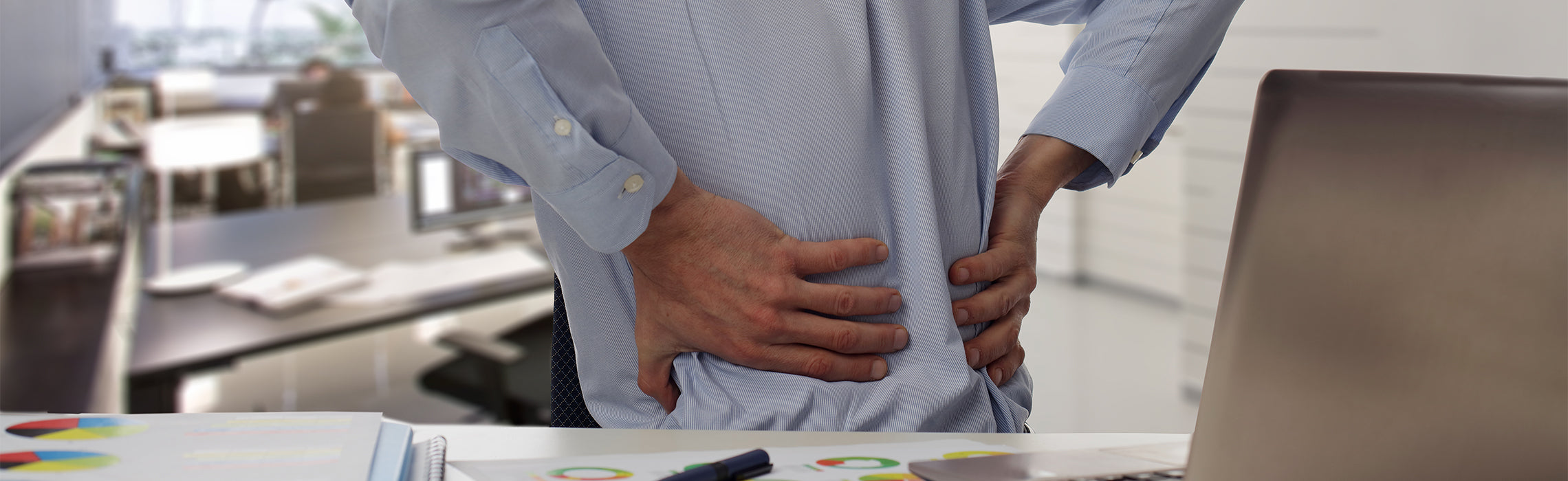 Person standing at an office desk holding the lower back with both hands, with a laptop, charts, and paperwork nearby, representing lower back pain, poor posture, and workplace strain.