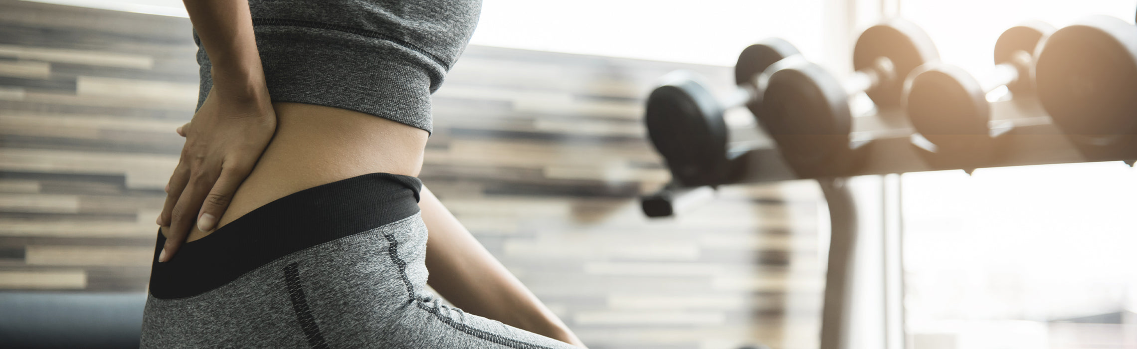 Close-up of a person in athletic wear sitting near gym equipment while holding the lower back with one hand, representing lower back pain, fitness-related strain, and exercise injury prevention.