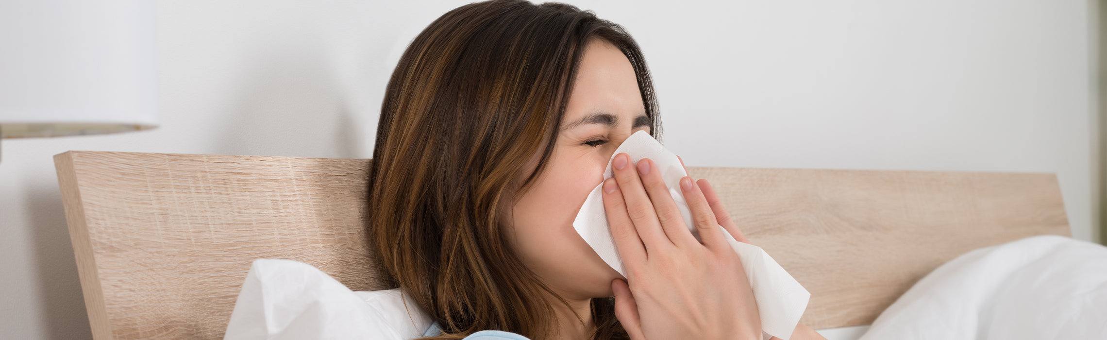 Woman sitting in bed blowing her nose into a tissue, appearing sick with cold or allergy symptoms.