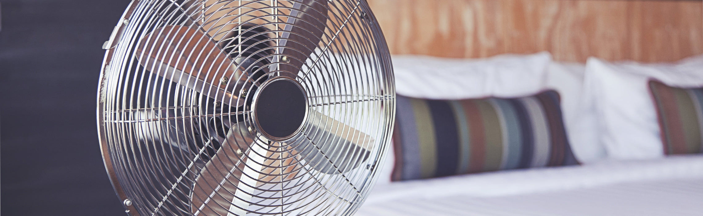 Electric fan positioned beside a neatly made bed in a bedroom setting.