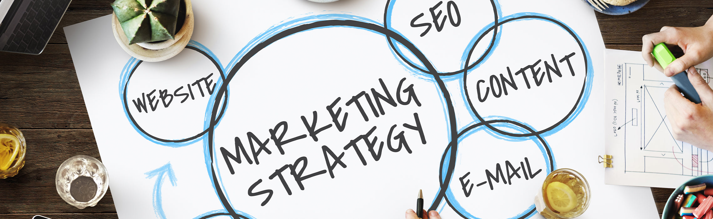 Marketing strategy diagram with hand-drawn circles labeled “MARKETING STRATEGY,” “SEO,” “CONTENT,” “E-MAIL,” and “WEBSITE” on a desk with a laptop, coffee, and notes, representing digital marketing planning.