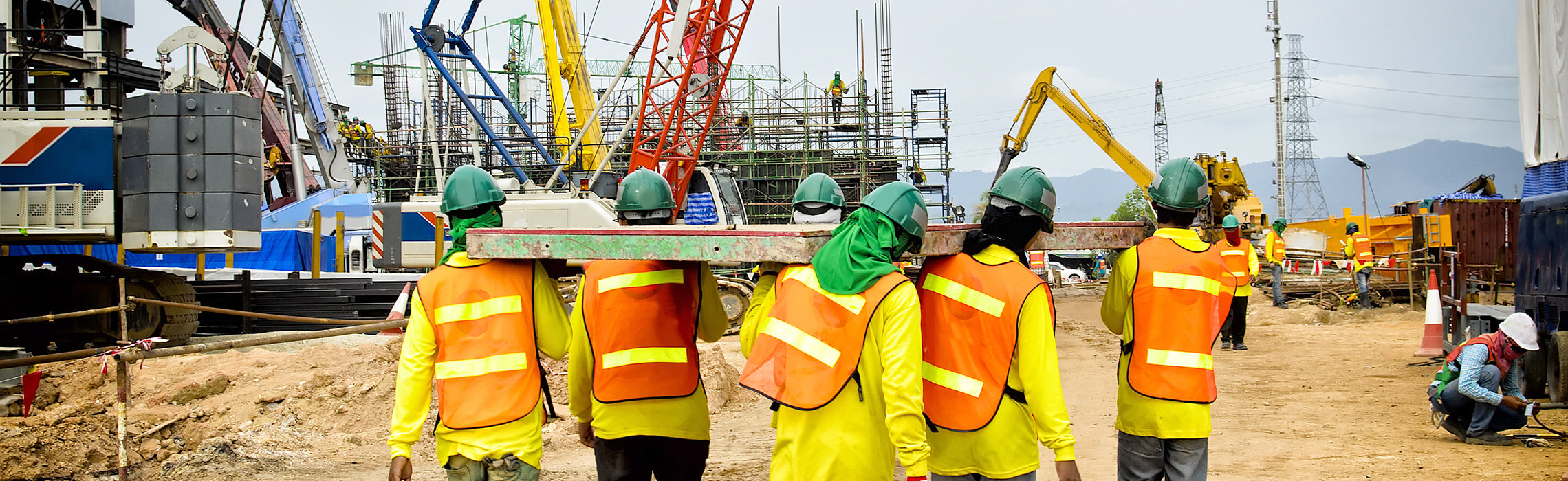Construction workers wearing green helmets and orange safety vests carrying a long beam across a busy construction site with cranes, scaffolding, and heavy machinery, representing teamwork, industrial work, and job site safety.