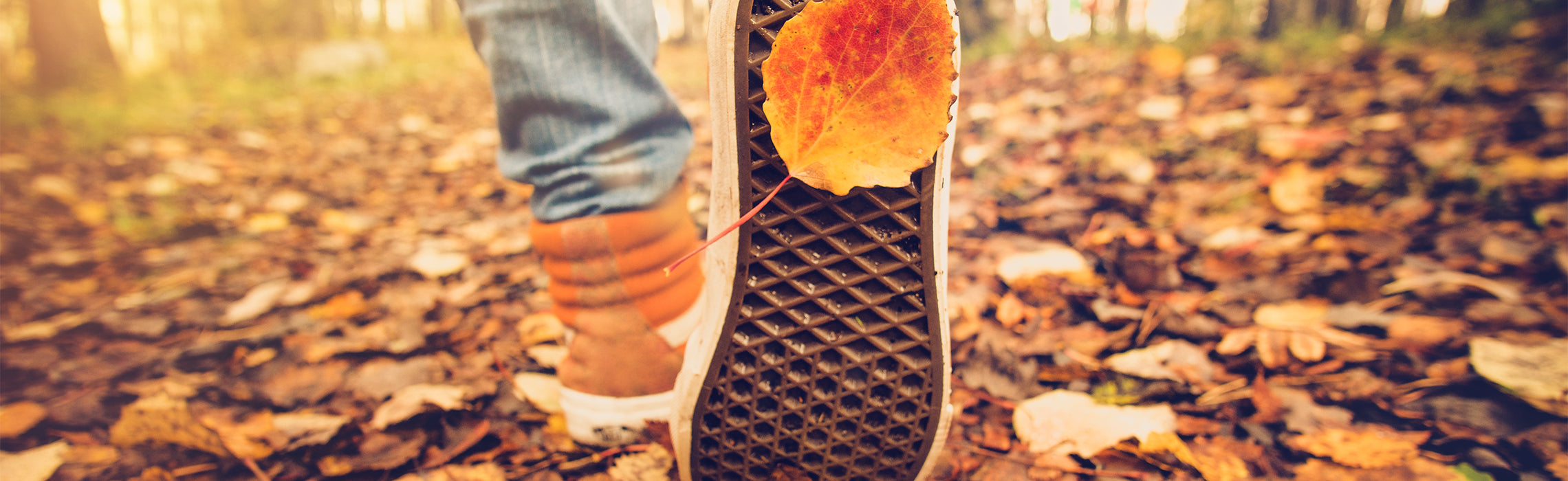 Close-up of a person walking outdoors on a leaf-covered path in autumn, showing the sole of a shoe with a highlighted heel area.
