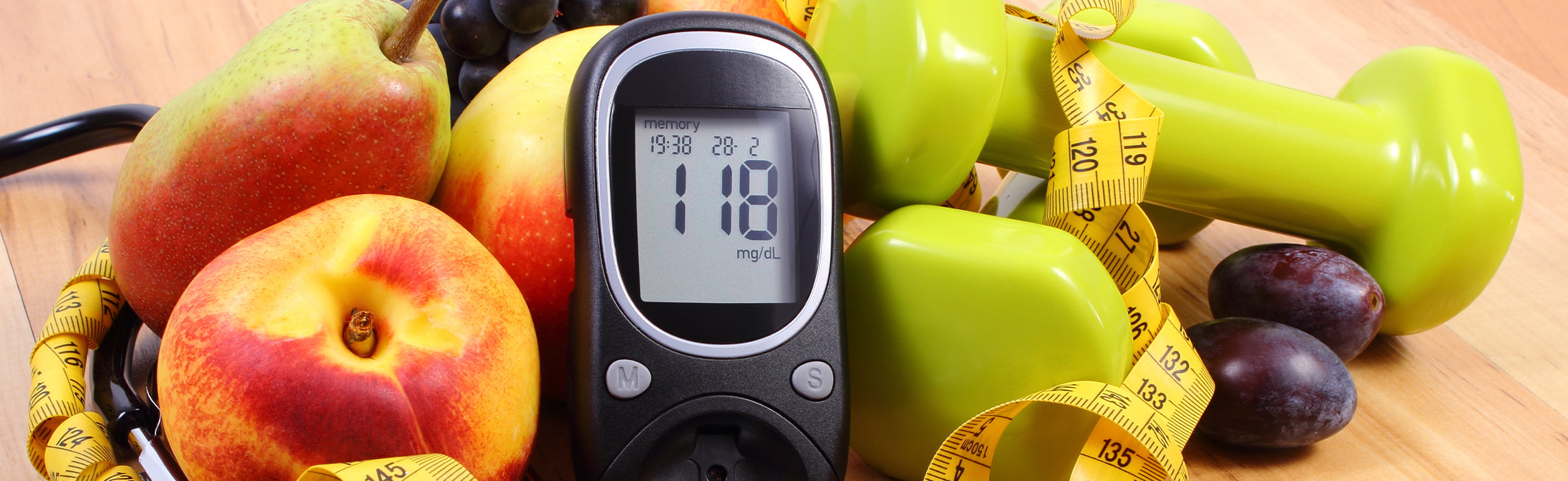 Digital glucose meter displaying a reading of 118 surrounded by fresh fruit, green dumbbells, and measuring tape on a wooden surface.