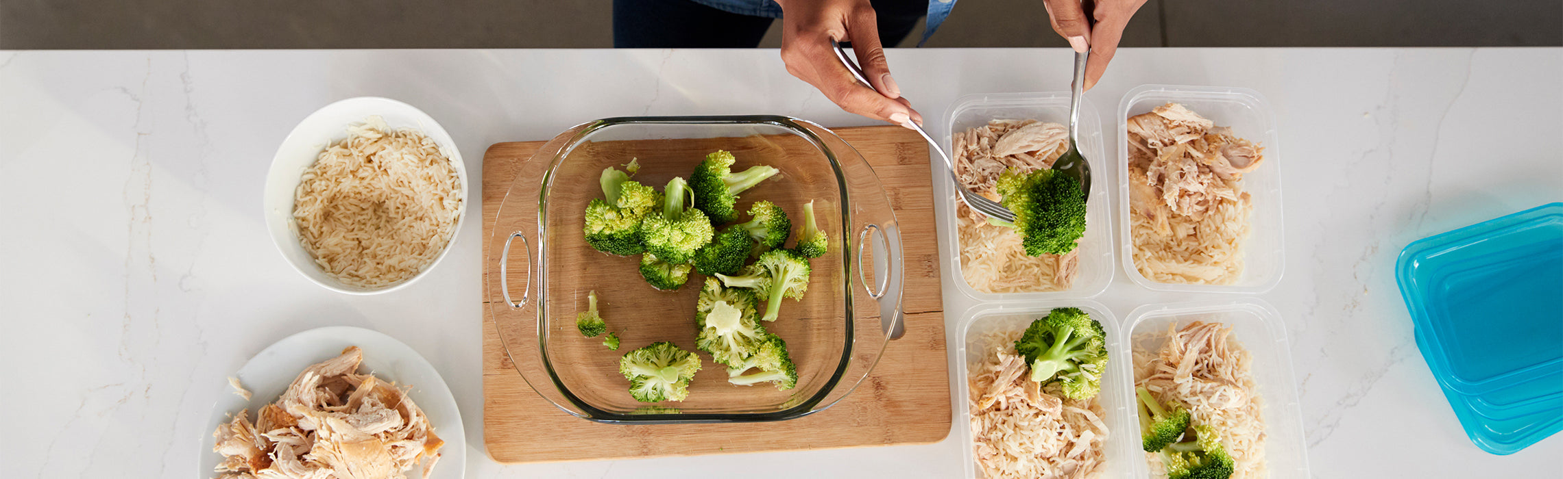 Top-down view of meal prep with broccoli, rice, and shredded chicken being portioned into clear containers on a kitchen counter. No visible text or branding.