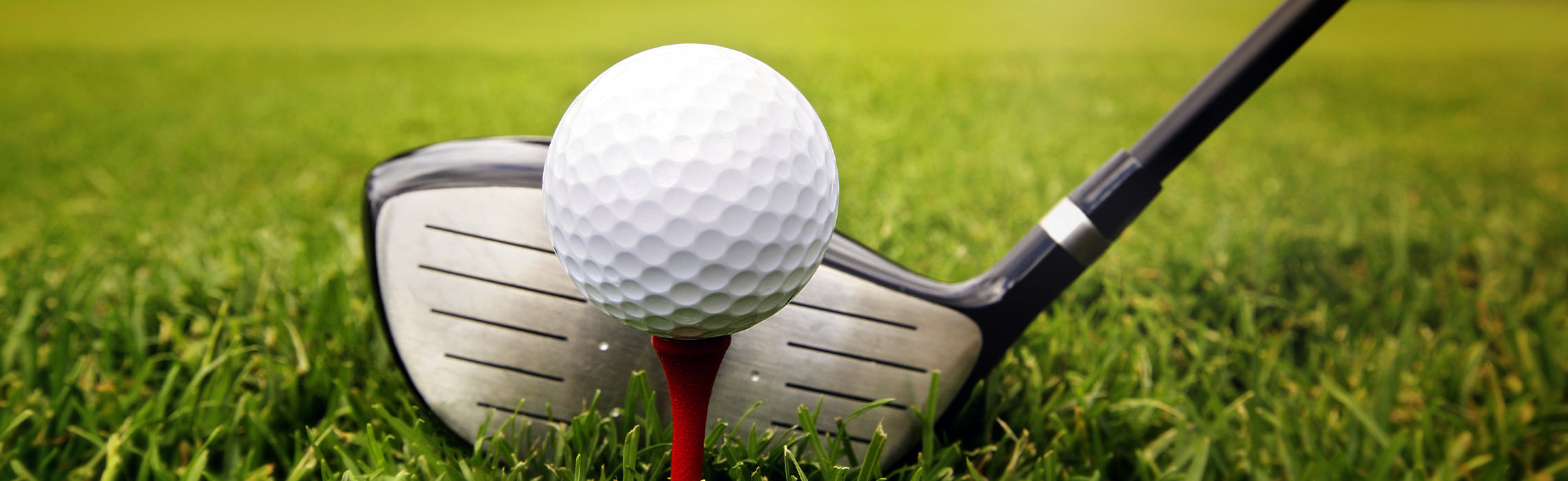 Close-up of a golf ball perched on a red tee in green grass with a metal golf club positioned behind it, representing golf practice, tee shot setup, and outdoor sport activity.