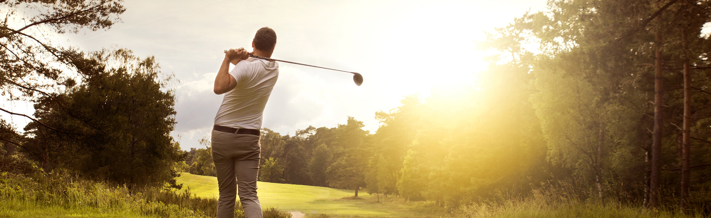 Person swinging a golf club on a sunlit course surrounded by trees, with bright sunlight in the background, representing golf, outdoor recreation, and leisure sports.