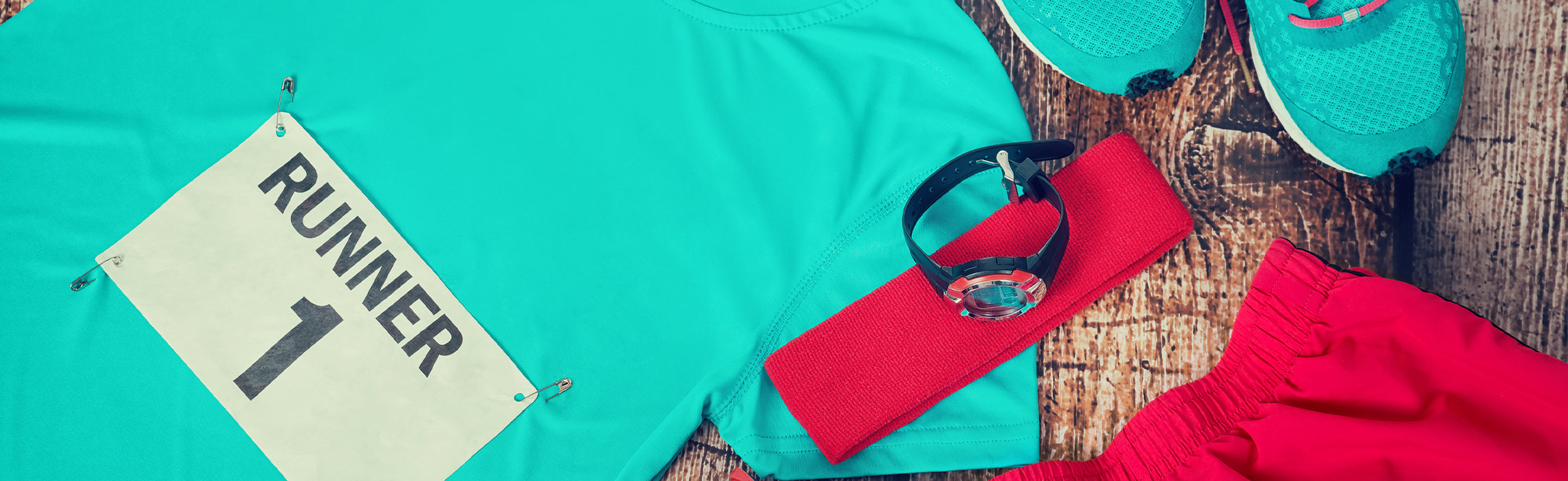 Flat lay of running gear including a teal athletic shirt, race bib labeled “RUNNER 1,” teal running shoes, red shorts, fitness band, and smartwatch on a wooden surface, representing race day preparation, fitness training, and active lifestyle.