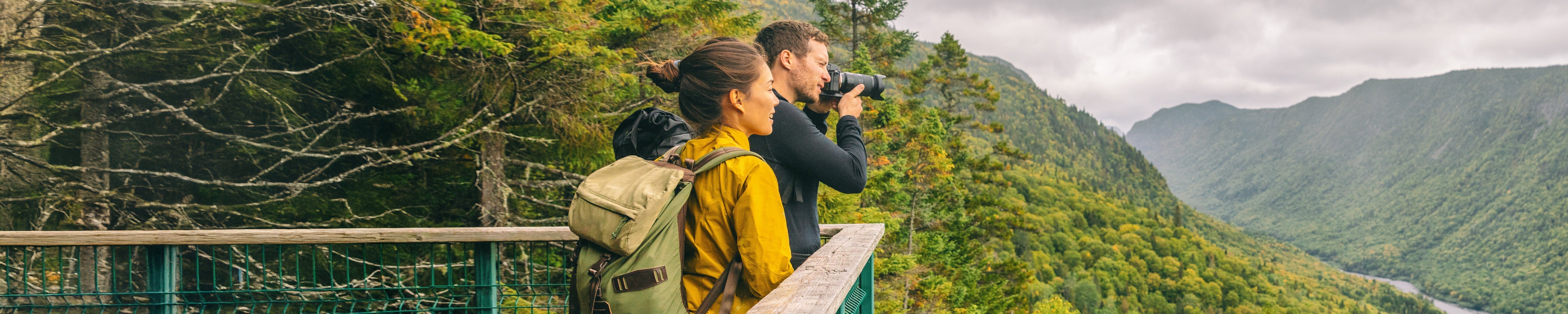 Two people with backpacks standing on a scenic overlook in a forested mountain area, one using binoculars; wooden railing and expansive valley views highlight outdoor hiking and nature exploration. No visible text.