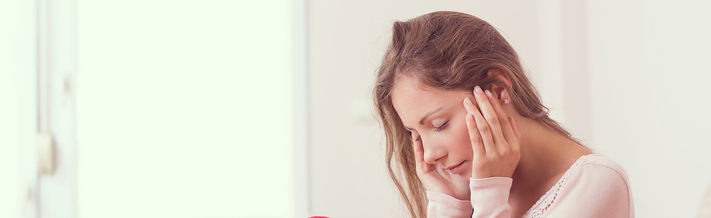 Close-up of a person sitting near a softly lit window, hands gently placed on both sides of the face against a neutral background. No visible text or branding in the image.