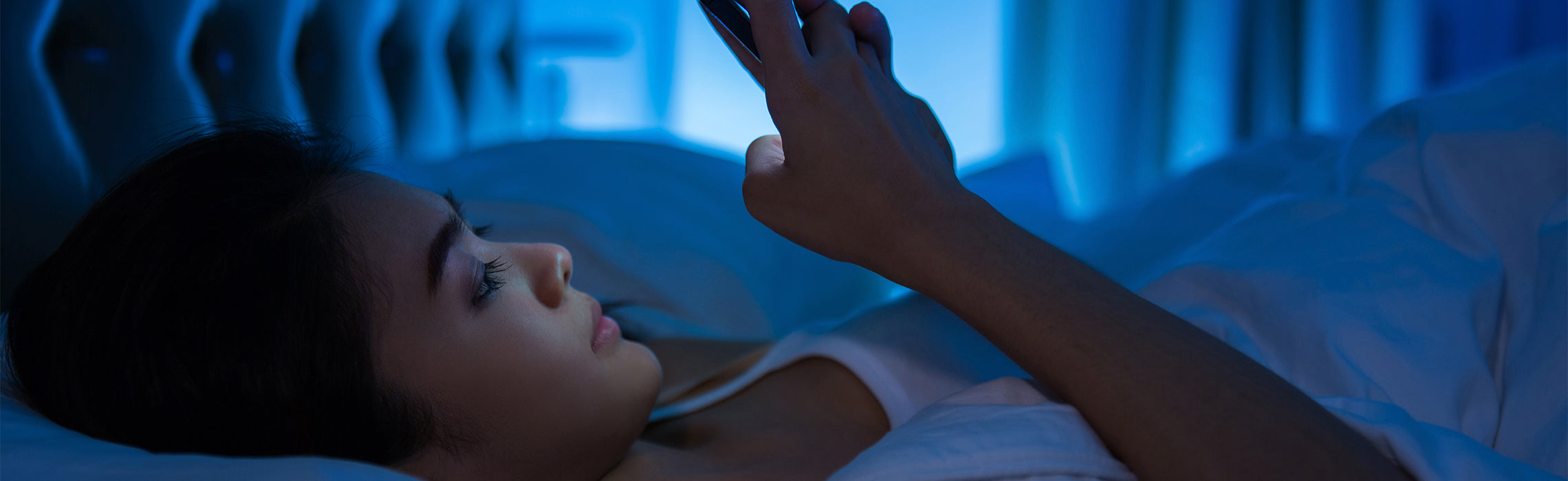 Person lying in bed at night using a smartphone, illuminated by blue light in a dark bedroom, representing screen time before sleep, insomnia, and disrupted sleep habits.