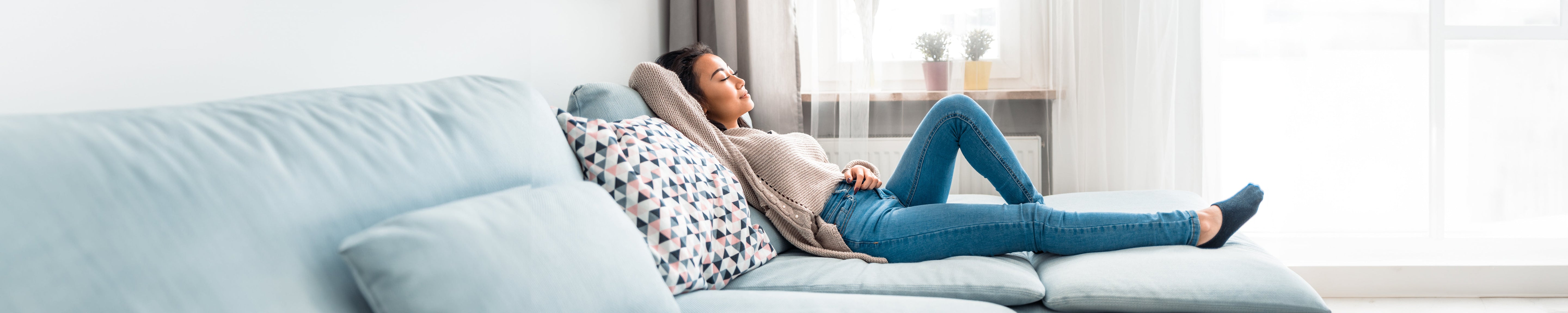 Person reclining on a light blue couch in a bright living room, legs stretched out and leaning against patterned pillows near a window with sheer curtains and potted plants, illustrating relaxation and comfortable home living. No visible text.