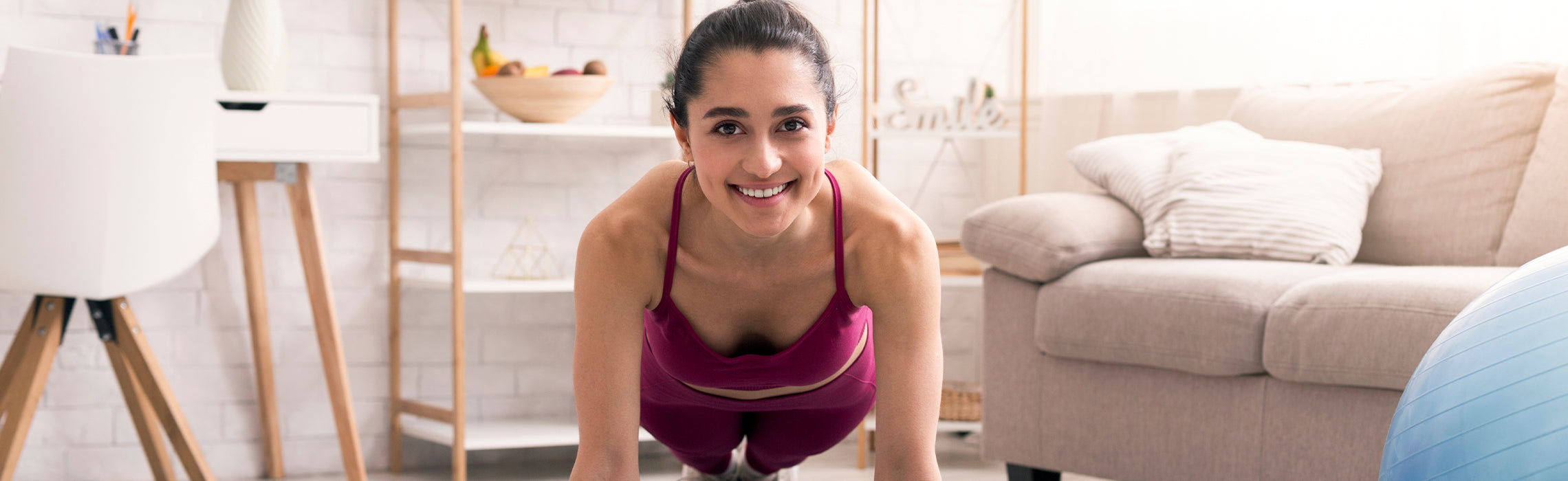 Person performing a plank exercise on a yoga mat in a bright living room, wearing athletic clothing, with a stability ball nearby, representing home workout, core strength, and fitness routine.