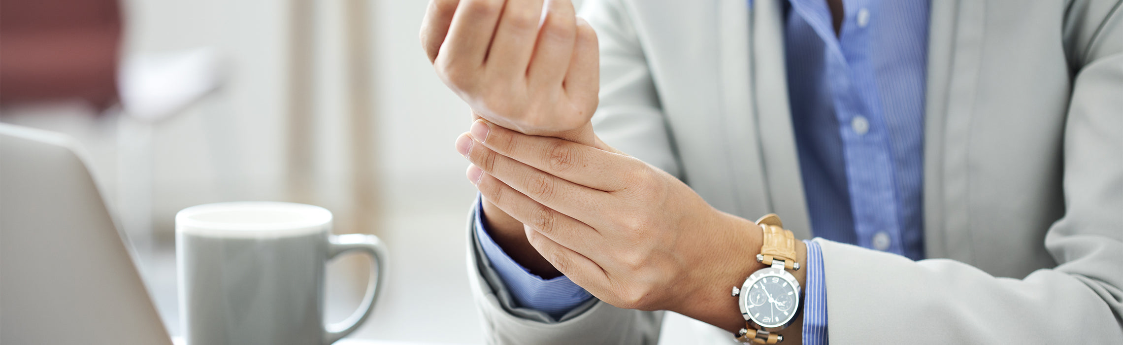 Close-up of a person seated at a desk holding and pressing the wrist with one hand, wearing a watch, with a coffee mug and laptop nearby, representing wrist pain, strain, and workplace discomfort.
