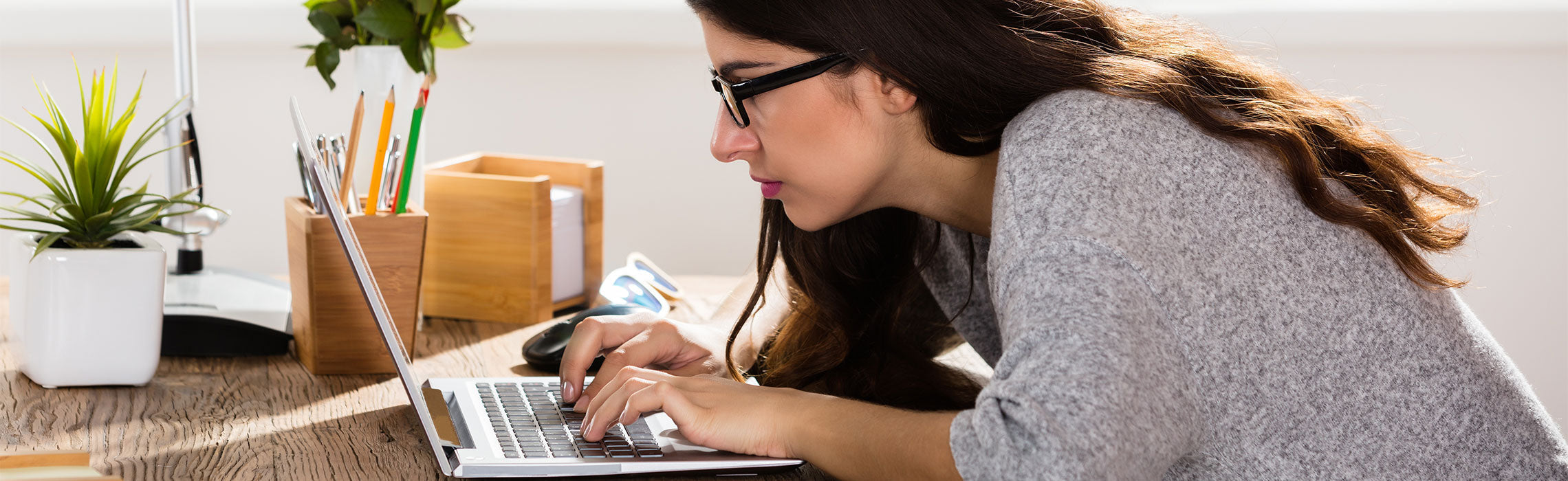 Person leaning forward while typing on a laptop at a desk, showing poor posture and potential neck or back strain. A wooden organizer with pencils and a potted plant sits nearby in a bright home office setting. No visible text or branding.