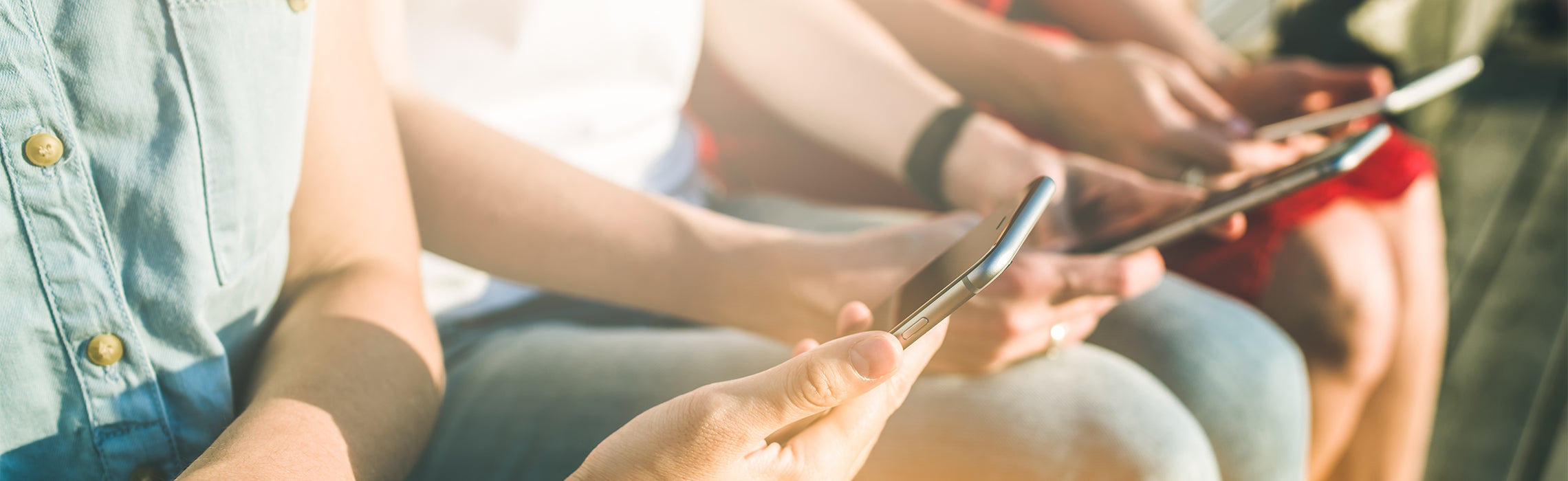 Close-up of several people sitting side by side outdoors, each holding and using a smartphone, representing mobile technology use, social media, and digital communication in everyday life.