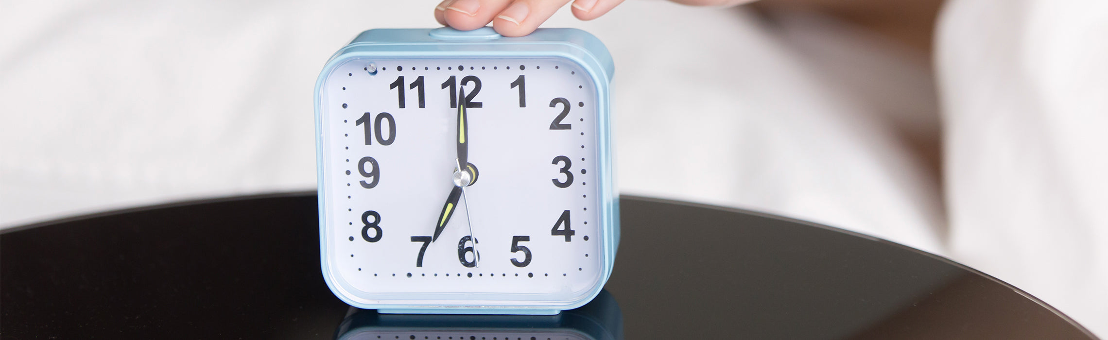 Close-up of a light blue analog alarm clock showing approximately 7:00, with a hand pressing the top button, representing waking up, morning routine, and time management.