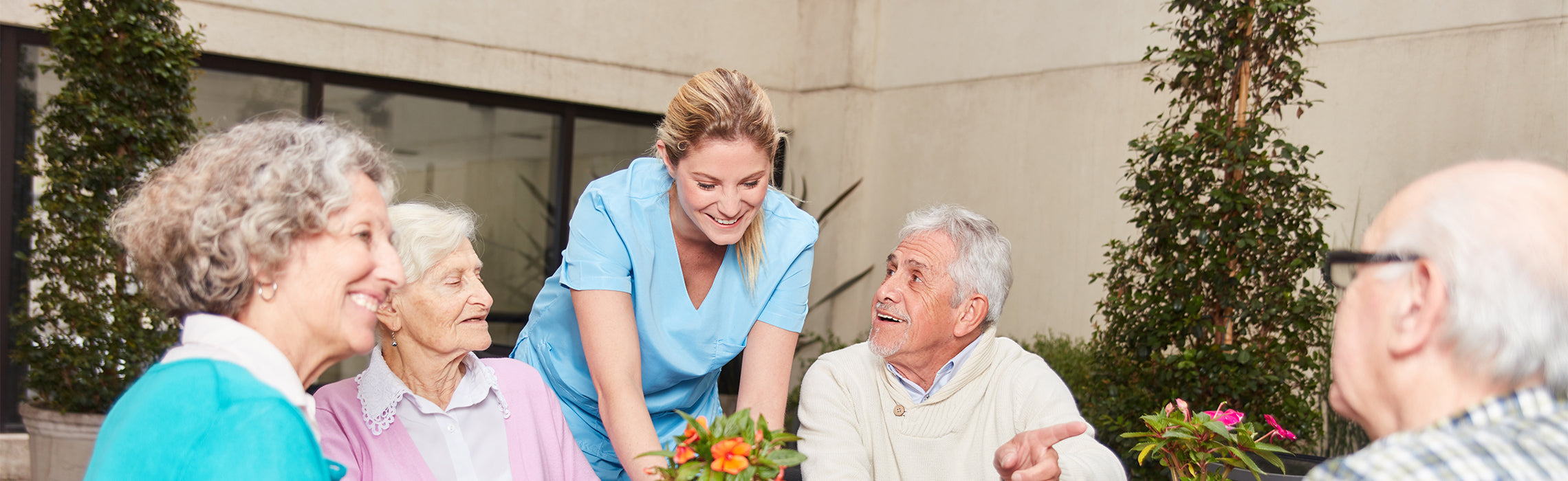 Healthcare professional in scrubs assisting older adults gathered around a table with plants in an outdoor patio setting, representing senior care, assisted living, and community support. No visible text.