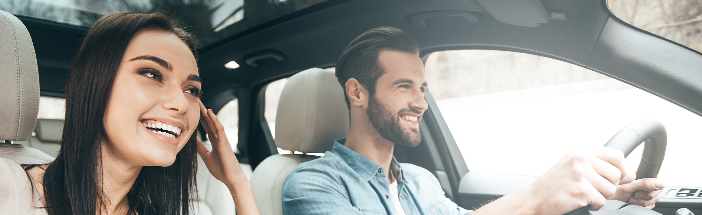Two people in a car during a daytime drive, with one driving and the passenger looking ahead in a bright, modern vehicle interior. No visible text or branding.