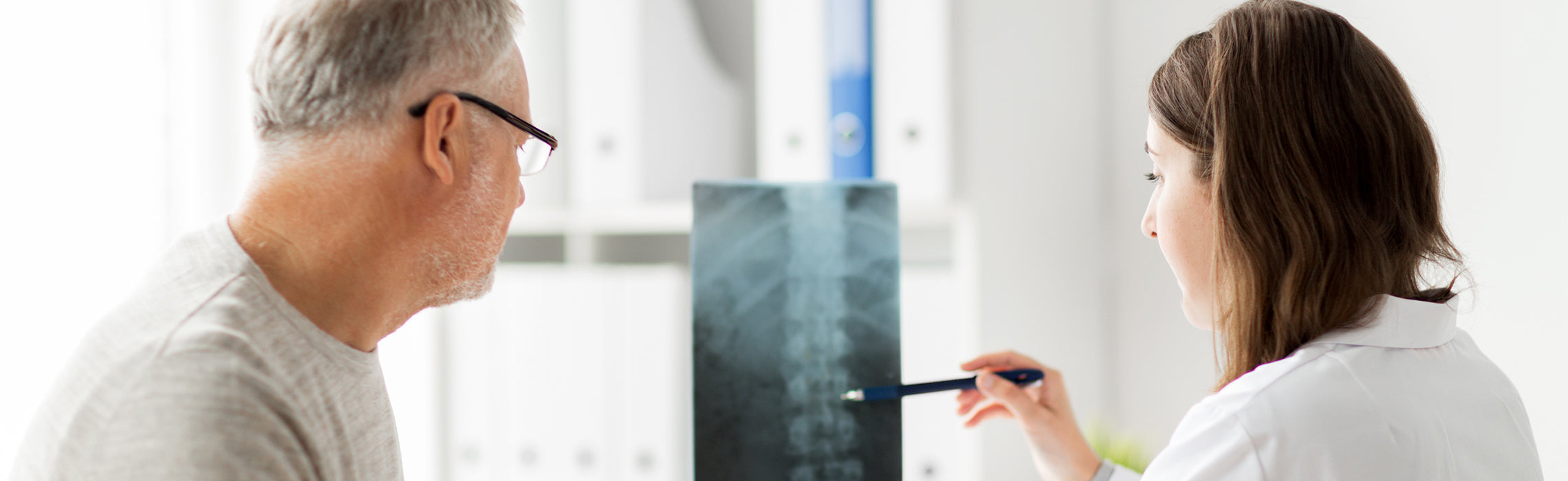 Healthcare professional showing a spine X‑ray to an older adult in a clinic, using a pen to point at the image, representing medical consultation, back health, and diagnostic care.