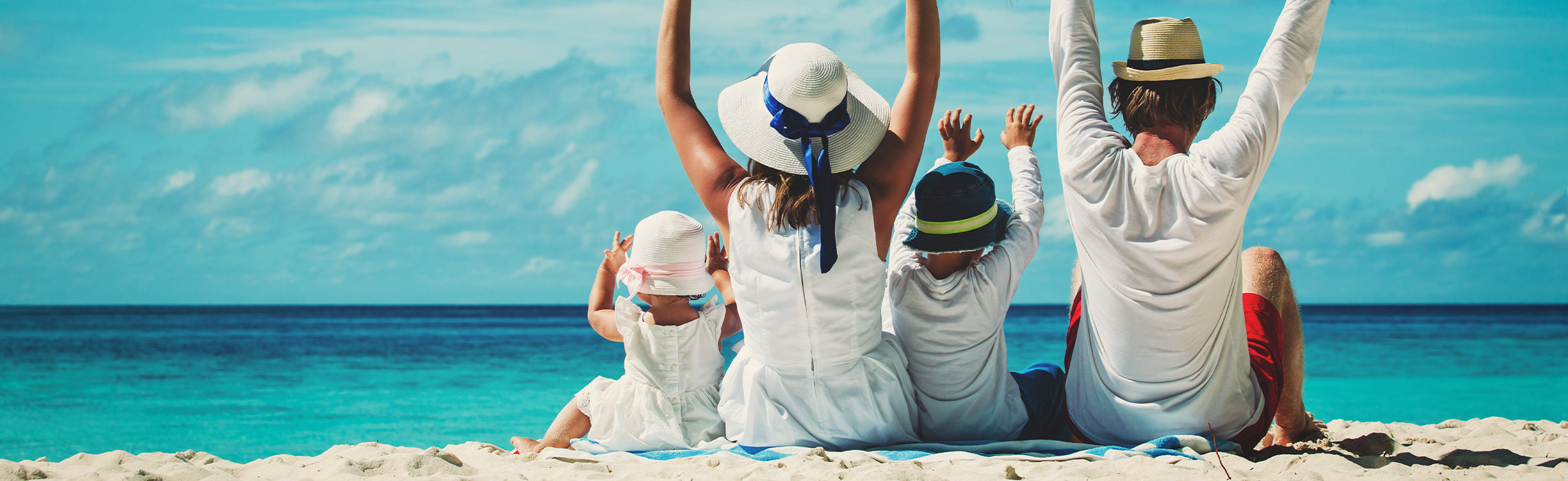 Family of four sitting on a beach with arms raised, facing the ocean, representing vacation, travel, and relaxation.