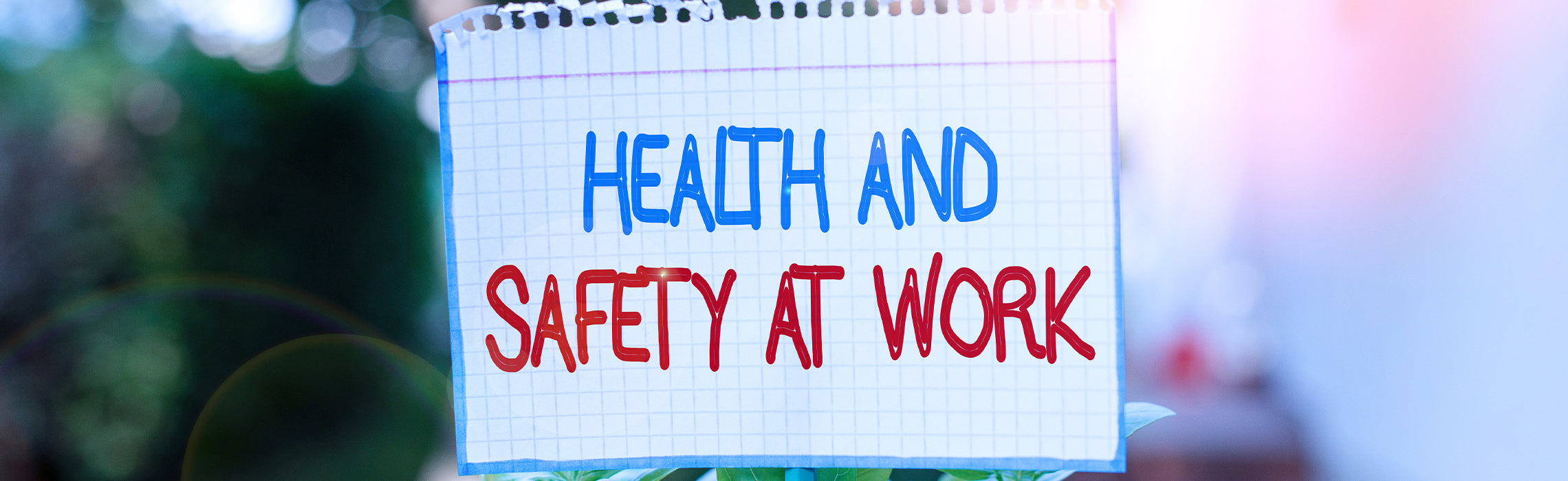 Handwritten sign on grid paper reading “HEALTH AND SAFETY AT WORK” in blue and red lettering, displayed outdoors with a blurred background, representing workplace safety awareness and health guidelines.