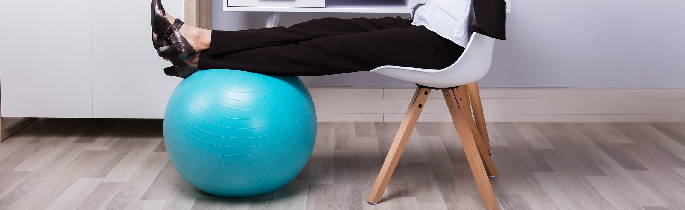 Person sitting in a modern chair with legs resting on a blue exercise ball under a desk, wearing work attire and dress shoes, representing active sitting, ergonomic workspace, and posture support in a home office environment.