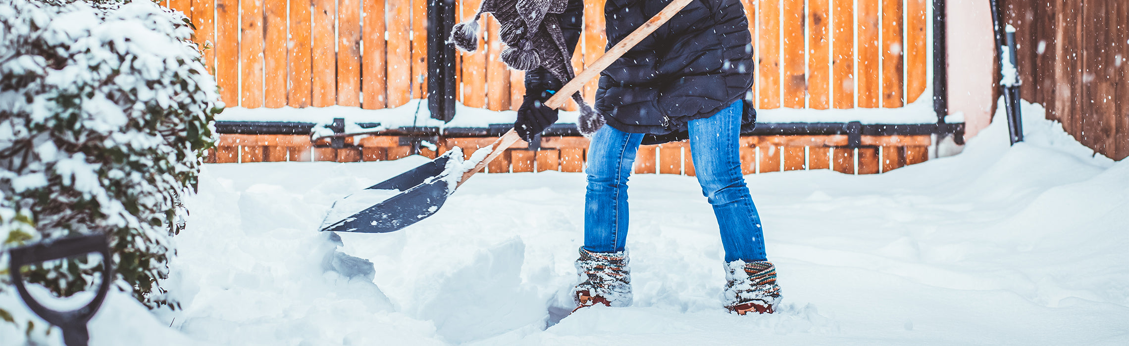 Person shoveling snow with a large snow shovel in a backyard during snowfall, wearing winter boots, jeans, and a jacket, with a wooden fence and snow-covered ground in the background, representing winter chores and outdoor activity.