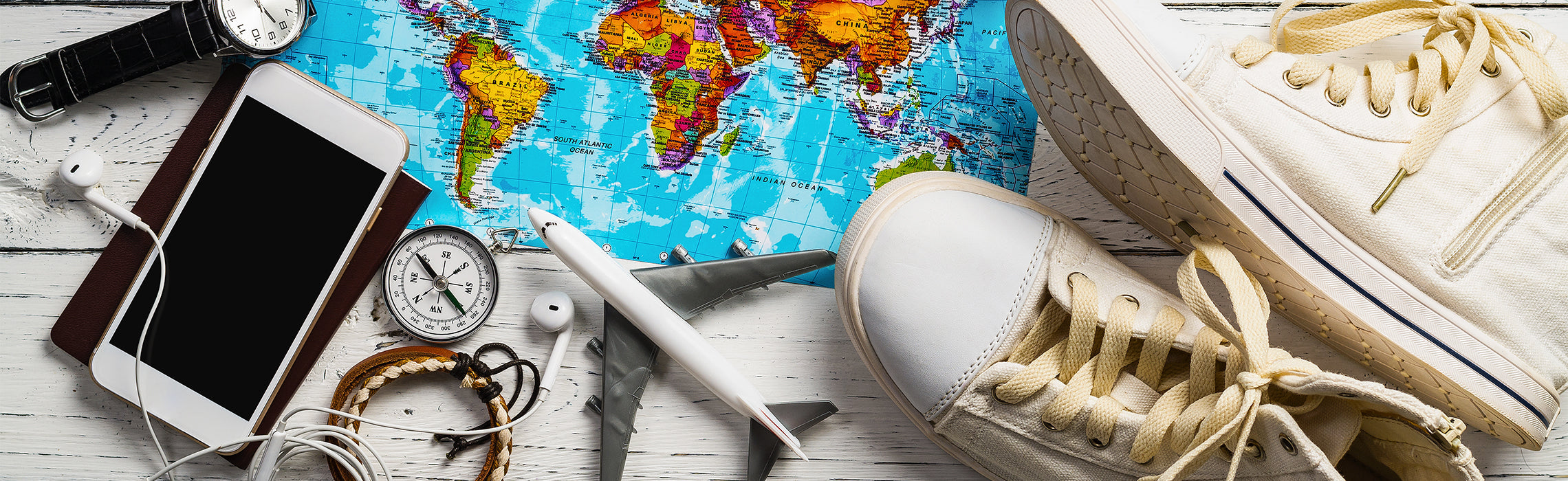 Flat lay of a world map with travel items including white sneakers, a model airplane, smartphone with earphones, compass, wristwatch, and bracelets on a wooden surface, representing travel planning, global exploration, and vacation essentials.