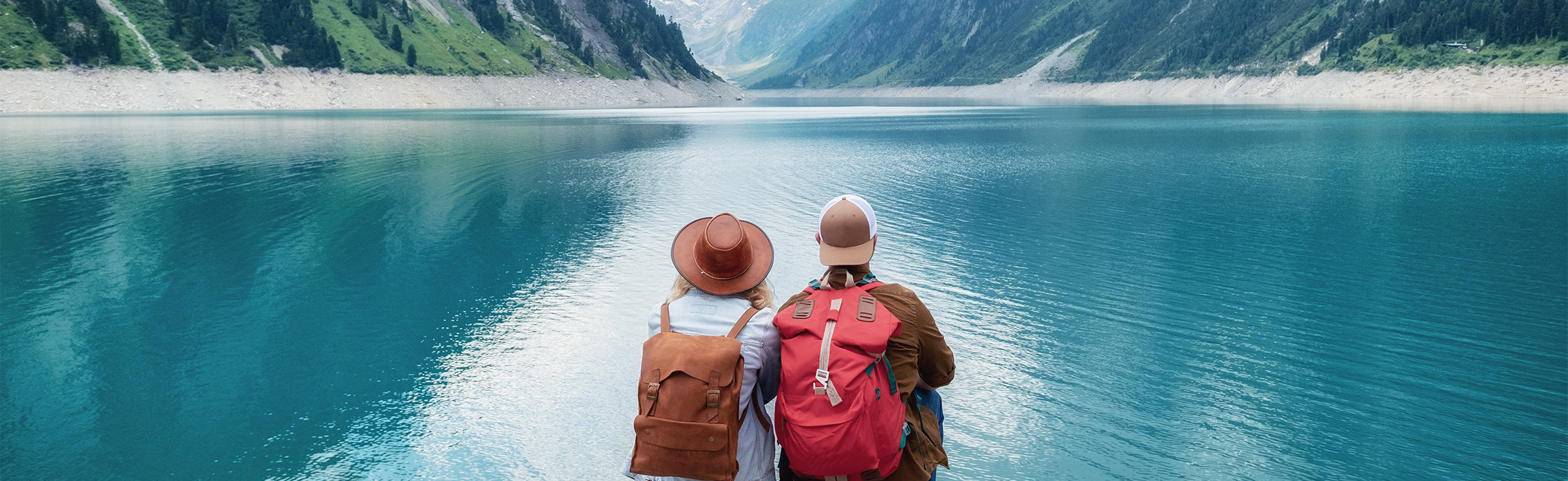 Two people with backpacks sitting side by side overlooking a calm turquoise mountain lake surrounded by steep green valleys, representing outdoor travel, hiking, and scenic nature exploration.
