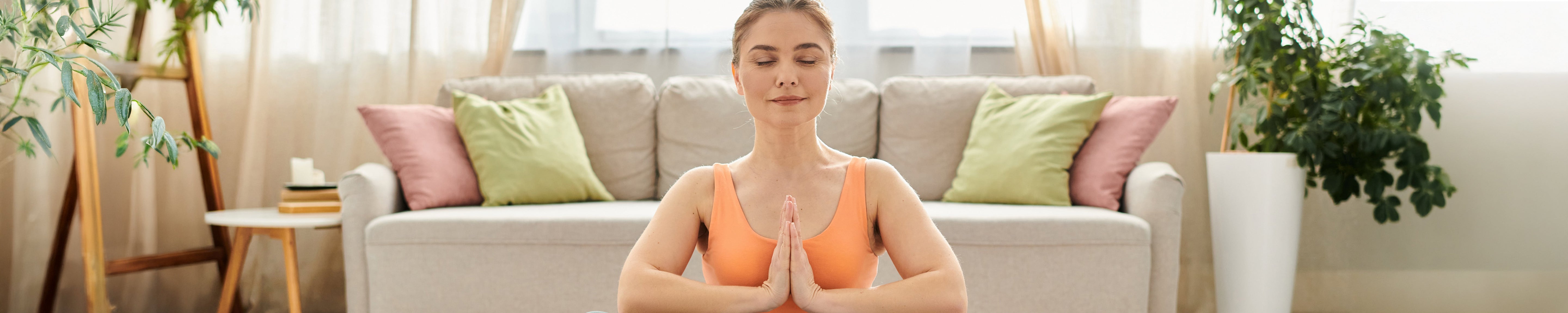 Woman in orange athletic wear practicing seated meditation with hands in prayer position in a bright living room.