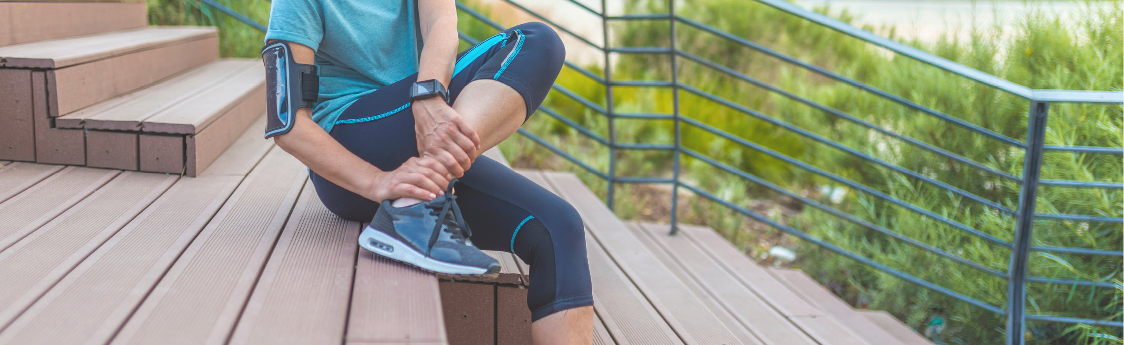 Person sitting on outdoor steps holding one ankle with both hands, wearing athletic clothing and running shoes, representing ankle pain, sports injury, and active lifestyle recovery.