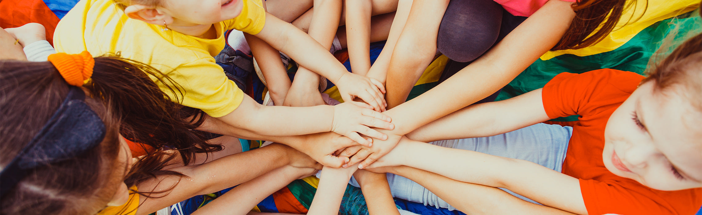 Top-down view of children placing their hands together in the center on a colorful parachute, representing teamwork, unity, and group play or youth activity.