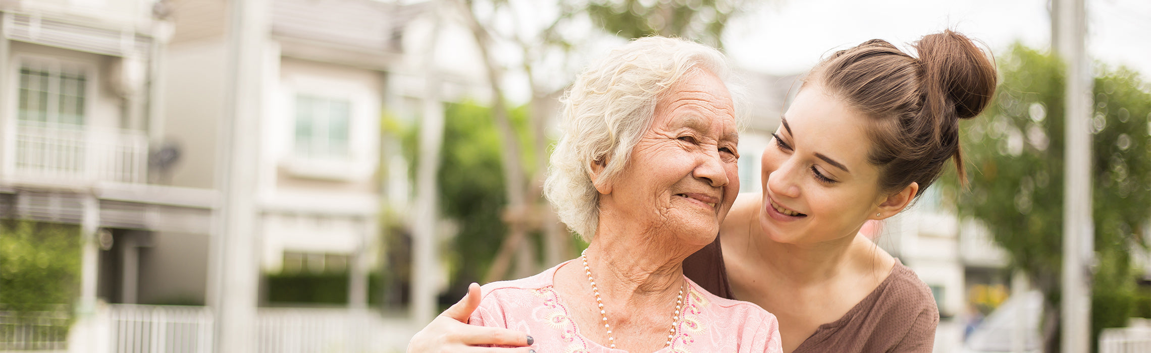 Older adult and younger person standing outdoors near residential homes, with the younger person’s arm around the older adult’s shoulders, representing caregiving, companionship, and senior support.