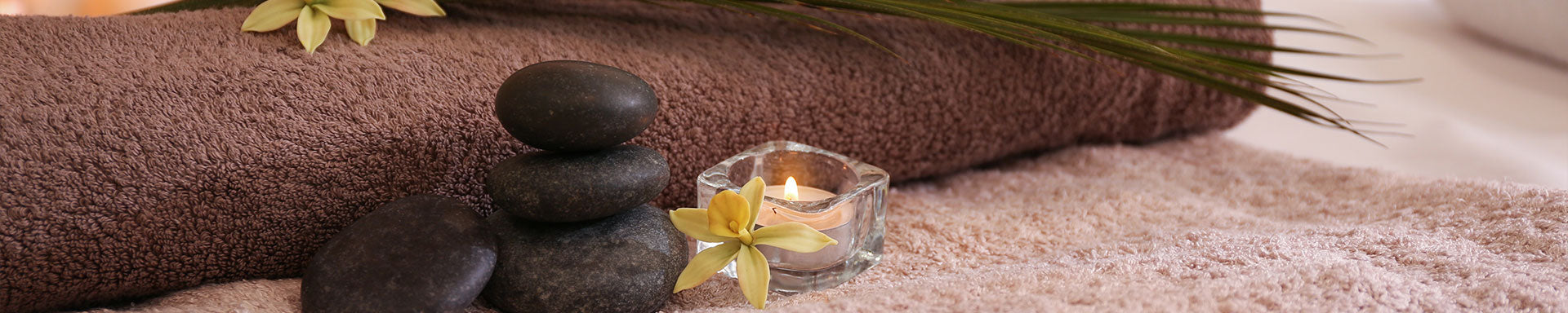 Horizontal view of a relaxation setup featuring a stack of smooth dark stones, a lit candle in a glass container, a small yellow flower, and rolled towels.