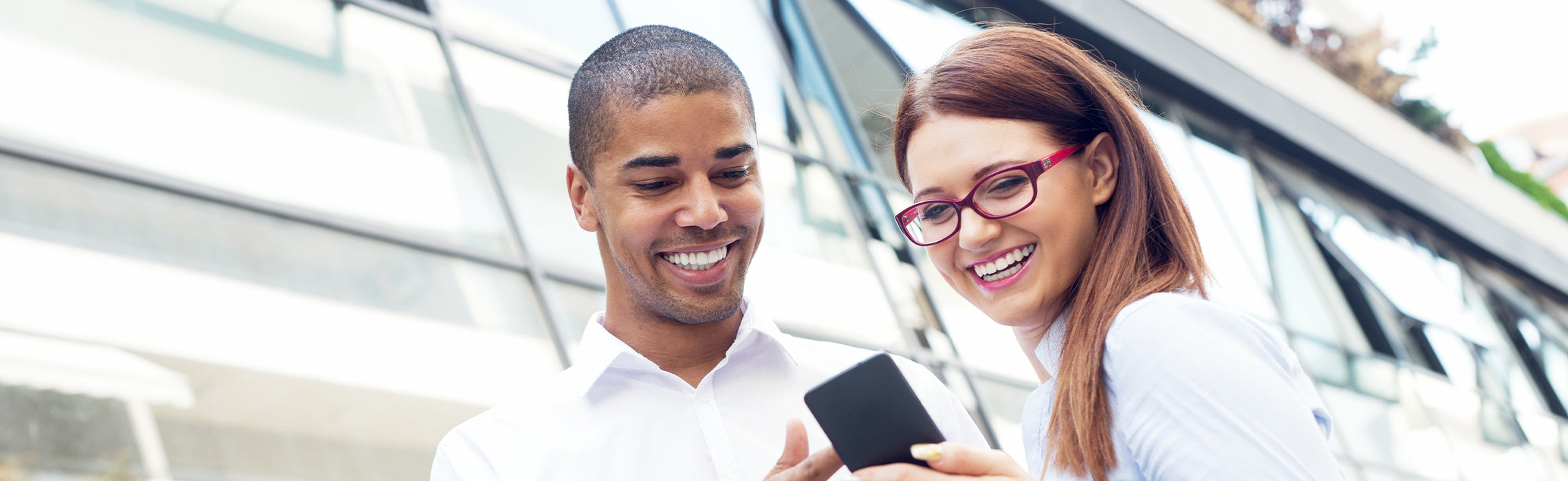 Two people standing outdoors near a modern glass building, smiling while looking at a smartphone together, representing mobile technology use, communication, and casual business interaction.